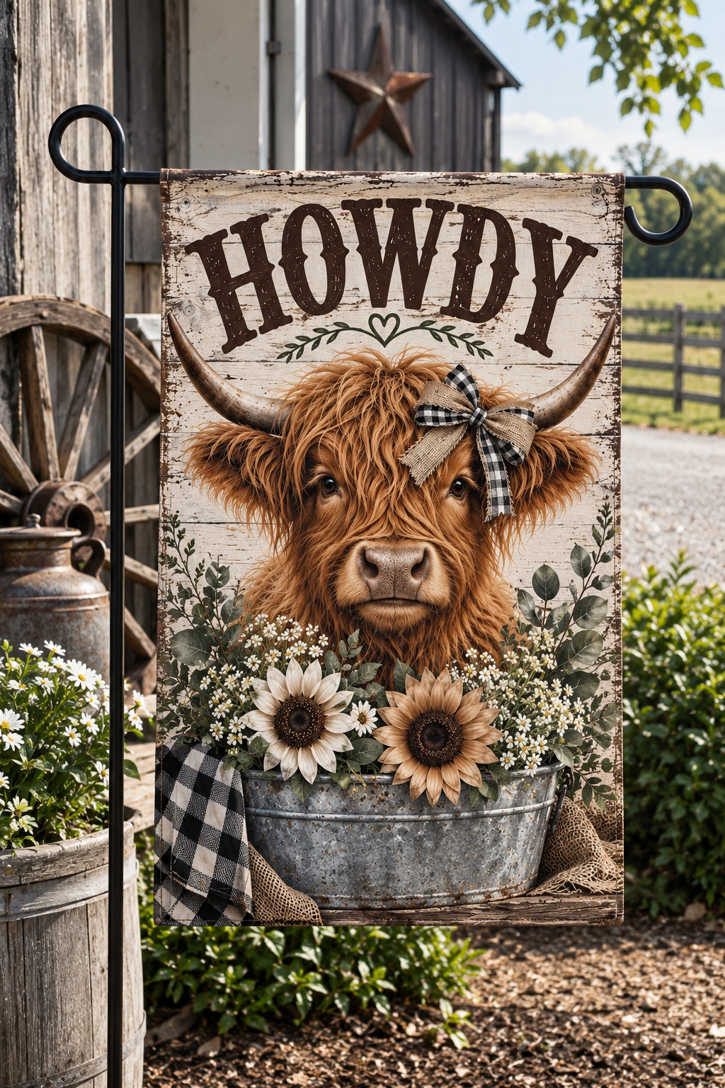 A decorative garden flag hanging on a black metal stand outdoors. The flag features a rustic wood-textured background with the word “HOWDY” at the top, a large brown highland cow in the center, sunflowers, white flowers, a galvanized metal tub, and a plaid bow.