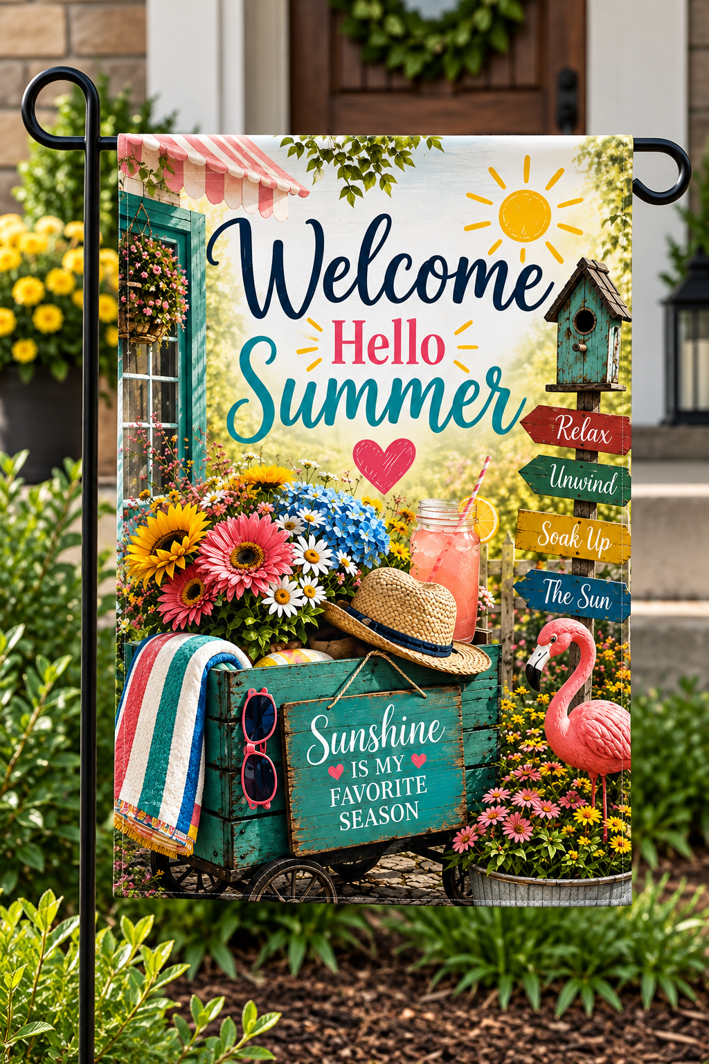 A decorative garden flag hanging on a black metal stand outdoors in front of a house door and garden plants. The flag features a colorful summer scene with the text 'Welcome Hello Summer,' flowers, a straw hat, a pink flamingo, a birdhouse, and signs reading phrases like 'Relax,' 'Unwind,' 'Seek Up The Sun,' and 'Sunshine IS MY FAVORITE SEASON.
