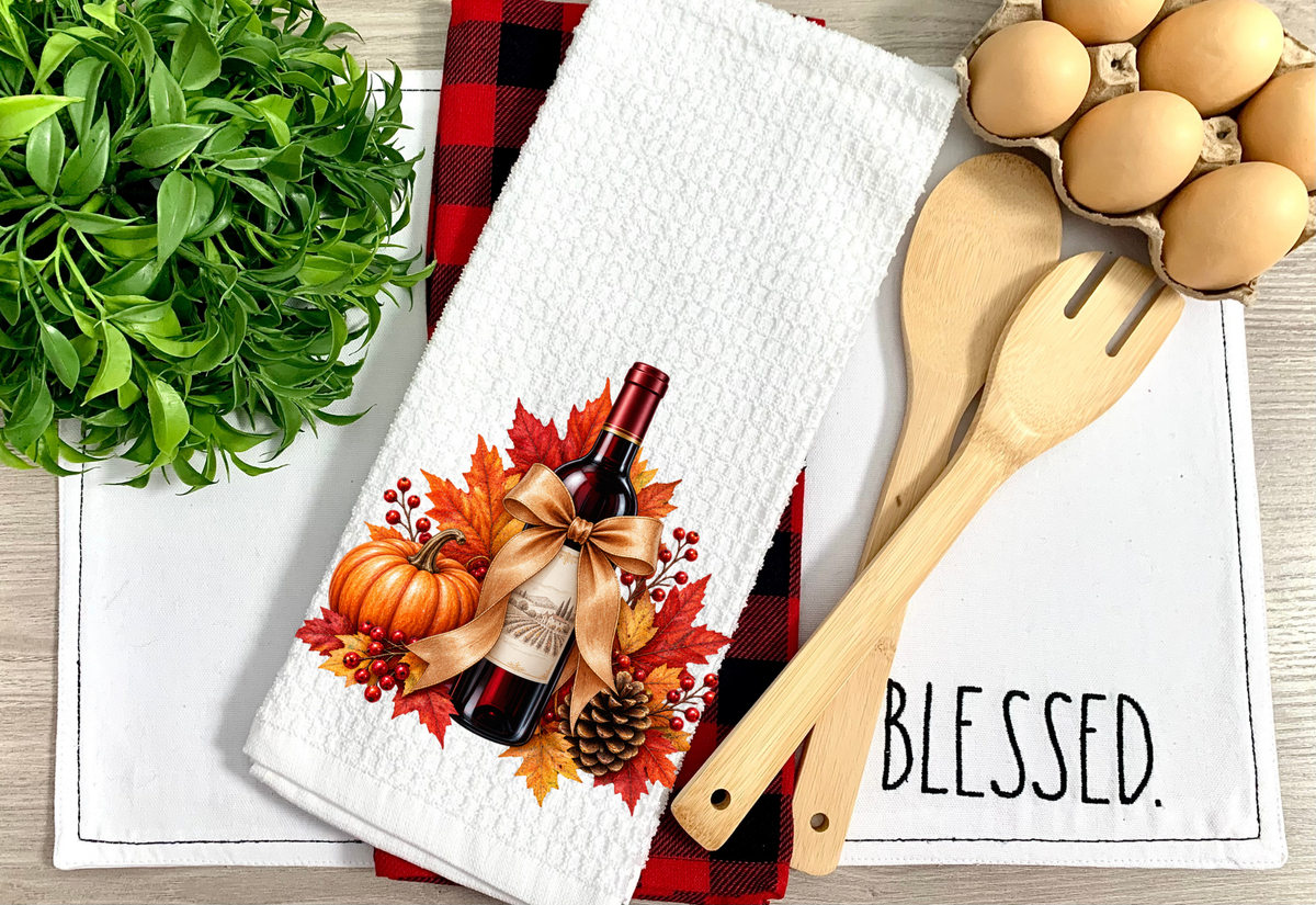 A white kitchen towel with a fall harvest design featuring a wine bottle, pumpkins, and autumn leaves, styled on a red and black buffalo plaid napkin with wooden utensils and a carton of eggs.