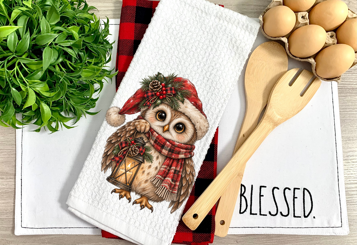 A white kitchen towel with a detailed illustration of a festive owl wearing a red plaid Santa hat and matching scarf, holding a lantern. The towel is styled in a flat-lay arrangement with a red and black buffalo plaid napkin, wooden kitchen utensils, a carton of eggs, and green foliage on a light wood background.