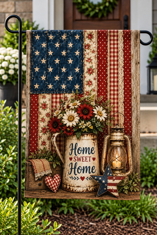 A decorative garden flag hanging on a black metal stand in a garden setting. The flag features a rustic American flag design with red, white, and blue panels, a vintage pitcher filled with flowers, a lantern, a small heart, a star, and the words 'Home Sweet Home' in script.